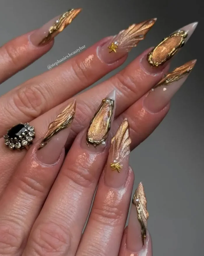 Long nude stiletto nails with intricate 3D metallic gold sculpted designs, including ridged textures and smooth framed elements, accented with pearls and gold stars.