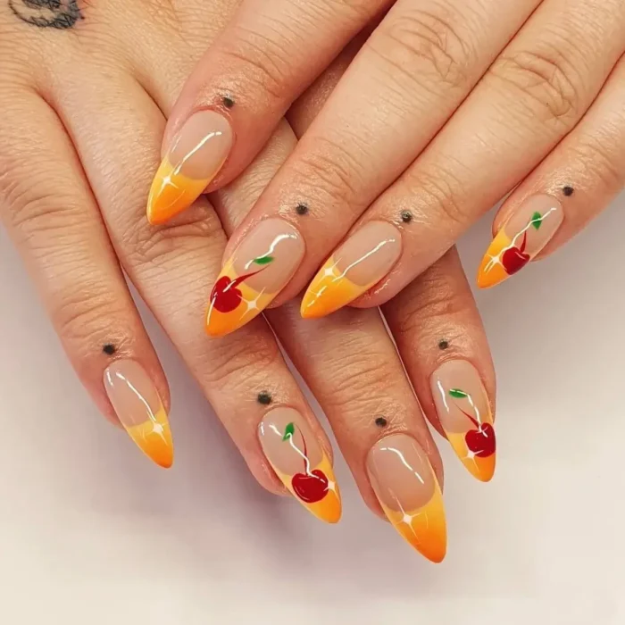 Medium stiletto nails with orange and yellow gradient tips, featuring hand-painted red cherries with green stems and small black dots near the cuticle.