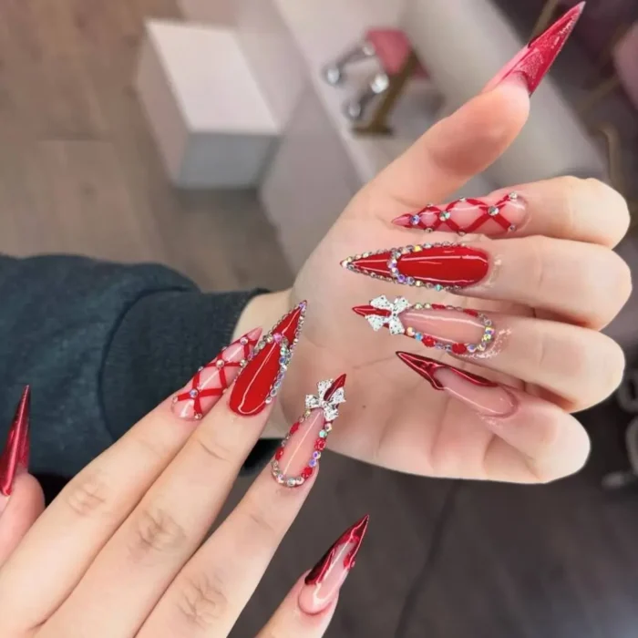 Long stiletto nails featuring a mix of solid ruby red, clear nails with red lattice patterns, and rhinestone embellishments including bows.