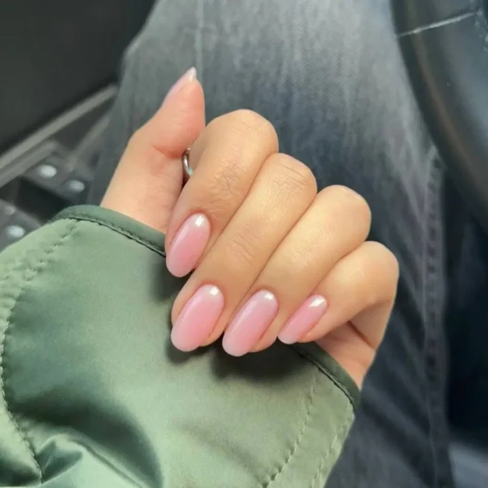 A hand with medium-length almond nails in a sheer pink polish, featuring a subtle white half-moon design at the nail base.