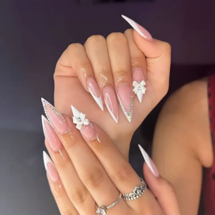 Long stiletto nails with white French tips, adorned with small white 3D bows and sparkling rhinestones along the sides.