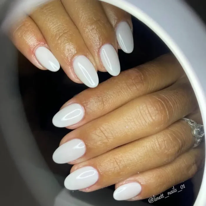 A set of hands with a flawless milky white manicure on medium-length oval nails, viewed through a ring light.
