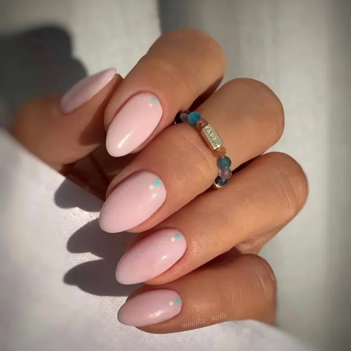 A hand with almond-shaped nails in a soft pink polish, with each nail featuring a tiny white and blue dot accent.