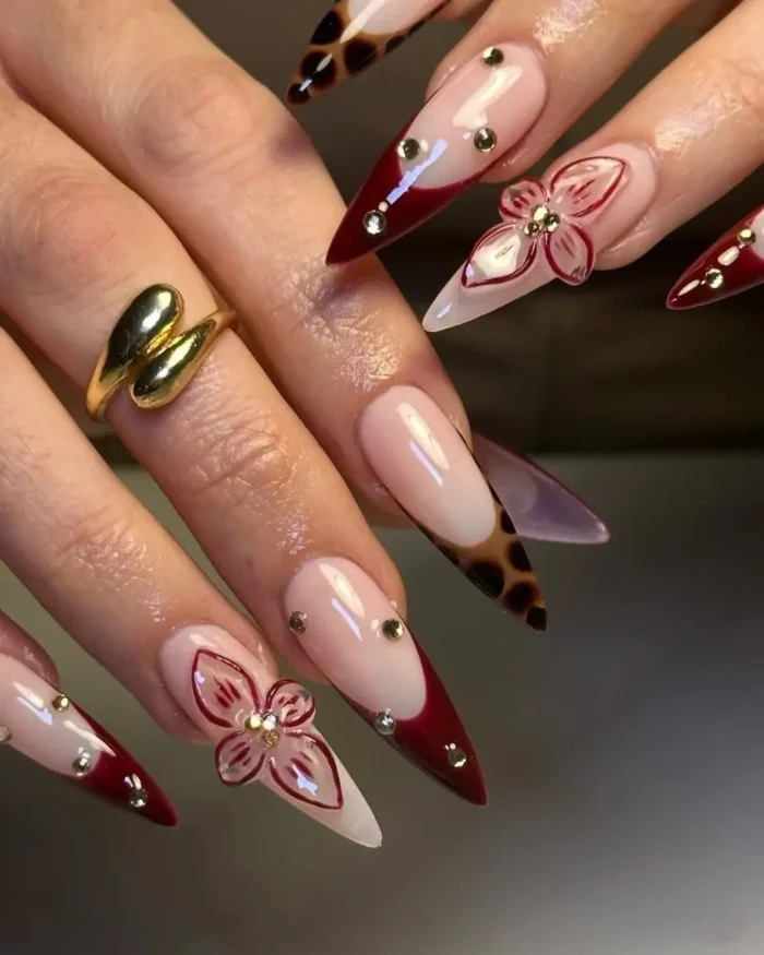 Long stiletto nails with burgundy tips, some featuring leopard print, and clear 3D flowers with gold studs.