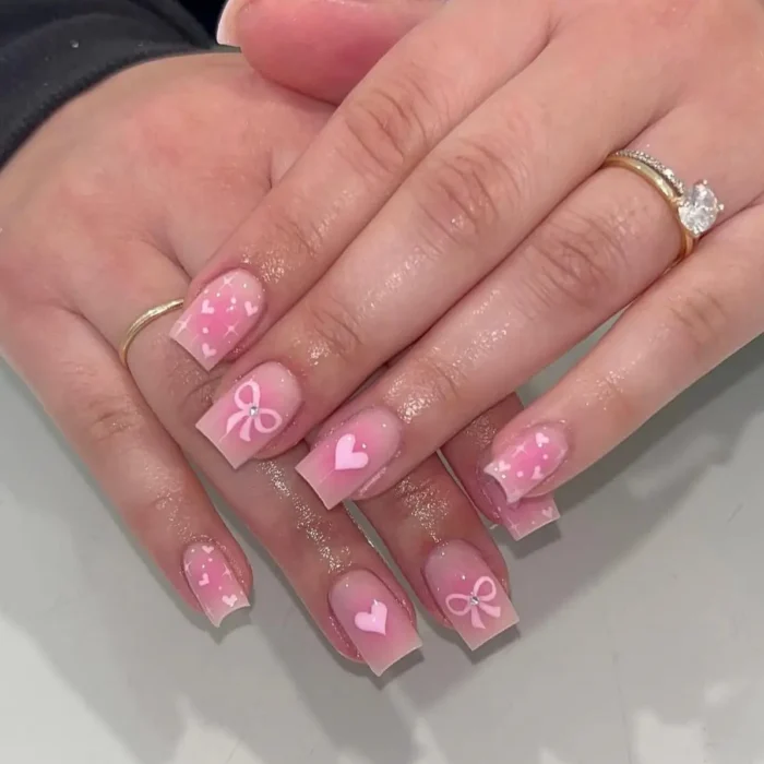 A manicure on square-shaped nails featuring a mix of pink hearts, bows, and glitter accents on a sheer pink base.