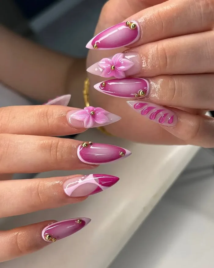Intricate pink stiletto nail design with a 3D flower, gold studs, and mixed art.