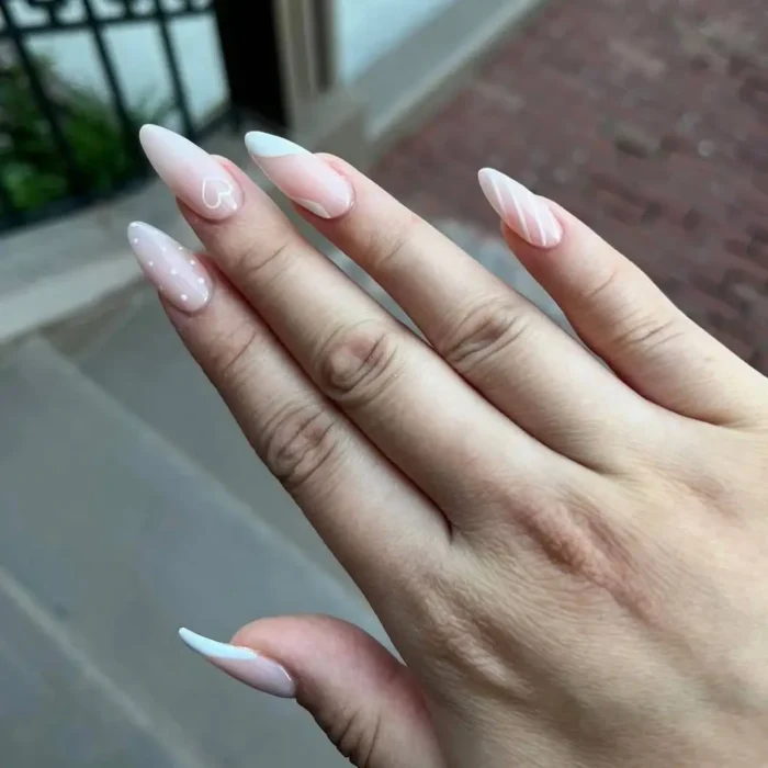 A hand with long almond nails showcasing a mix of simple white nail art, including hearts, dots, and stripes.