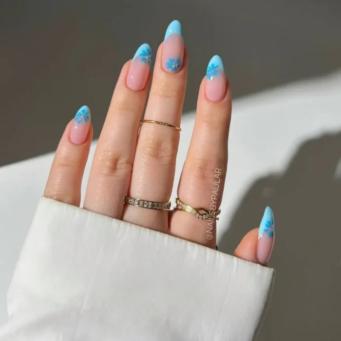 A close-up of almond nails with a sky blue french tip, accented with a small 3D flower.