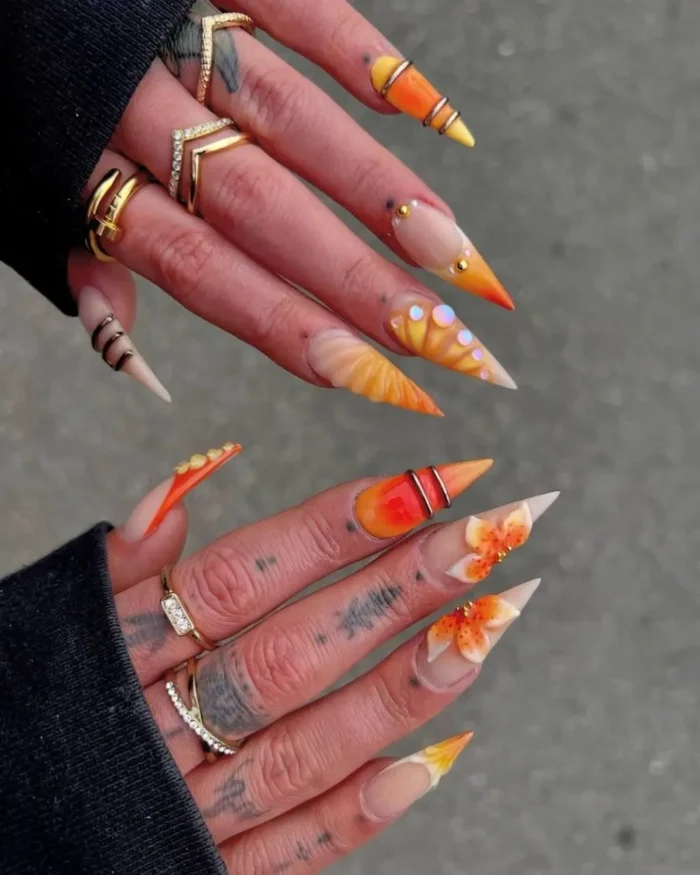 Long stiletto nails with an orange and yellow gradient, some featuring 3D flowers, rhinestones, and metallic ring accents.