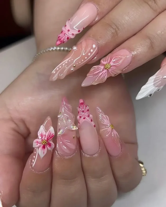Elaborate pink stiletto nail design with 3D flowers, a butterfly charm, and gold studs.