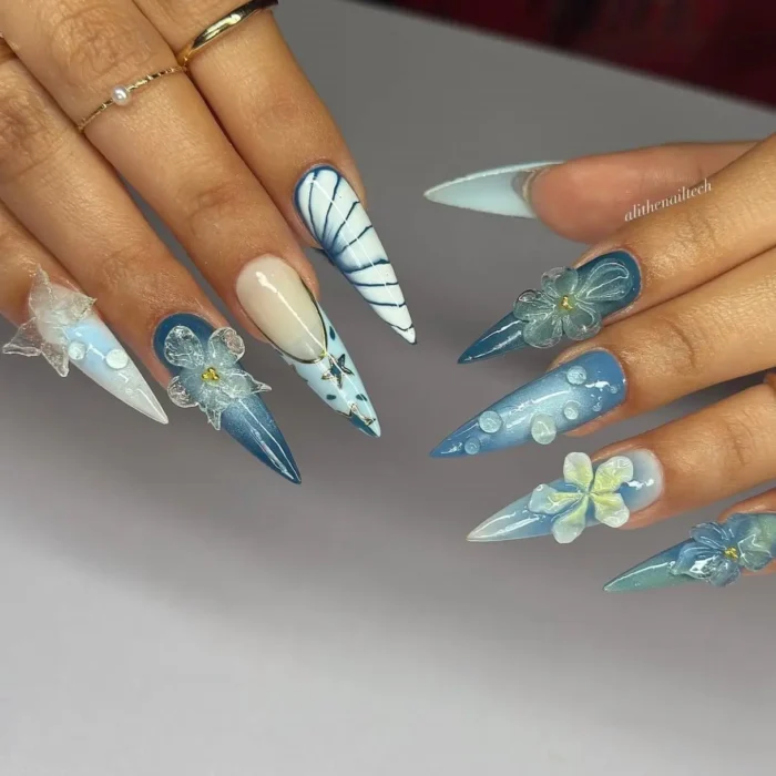 Long stiletto nails in shades of blue and white, adorned with 3D flowers, pearl-like dots, and wave patterns.