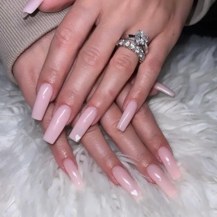 A manicure with long, square-shaped nails in a sheer pink, with two accent nails featuring a tiny, simple white heart.