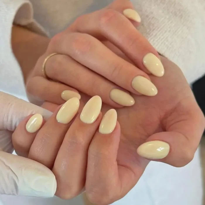 A simple oval nail design featuring a soft, buttery yellow polish on a pair of hands.