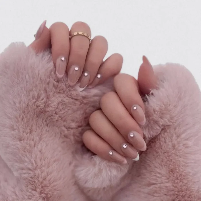 A close-up of long almond nails with a sheer pink base and a single delicate rhinestone accent near the cuticle.