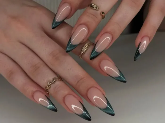 A close-up of two hands with long, almond-shaped nails featuring a dark emerald green cat eye french tip manicure, accented with delicate gold rings.