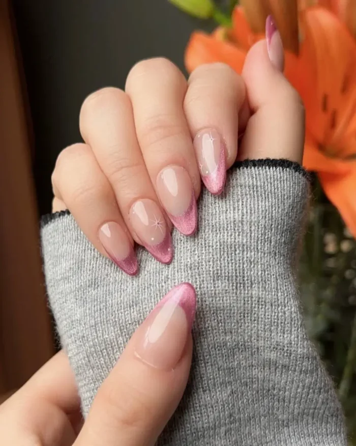 A hand resting on a grey sweater sleeve, showcasing a pink glitter cat eye french tip manicure on almond-shaped nails with subtle sparkle accents.
