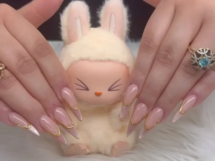 A pair of hands showing an elegant pink stiletto nail design with gold chrome frames and glitter tips, held in front of a small, cute figurine.