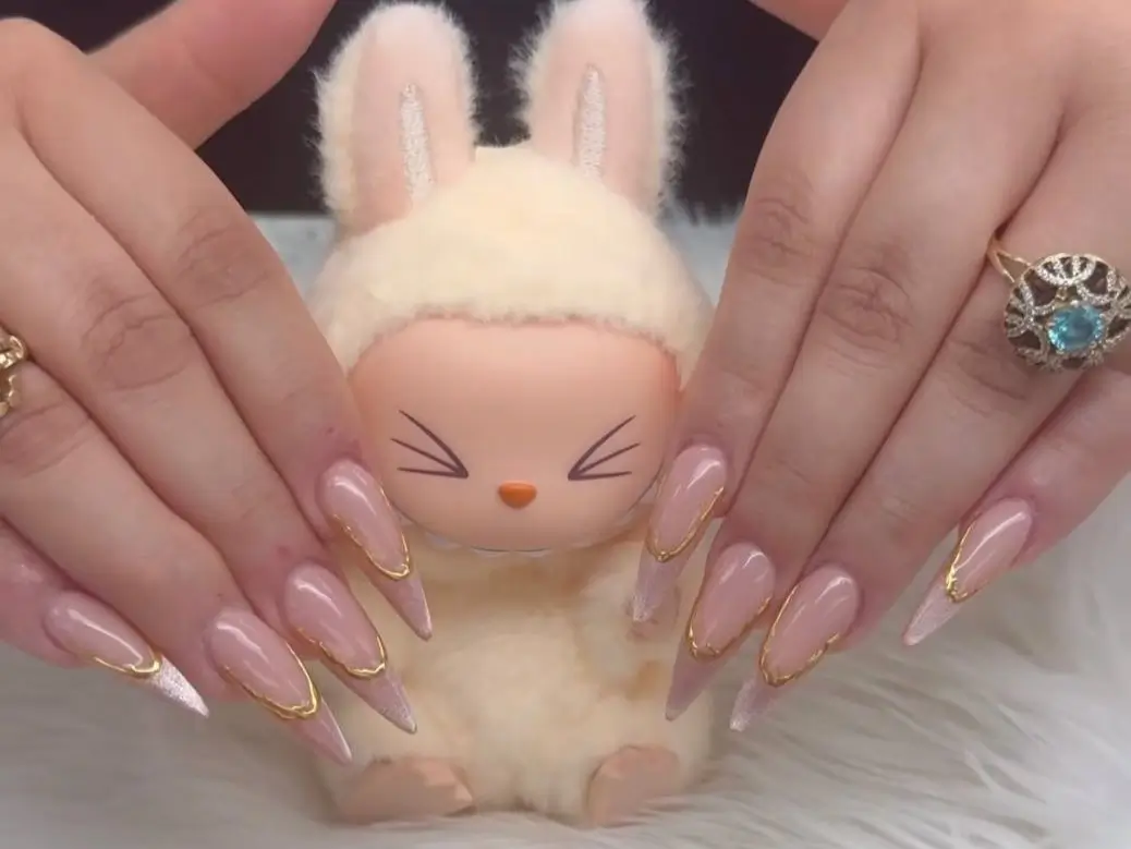 A pair of hands showing an elegant pink stiletto nail design with gold chrome frames and glitter tips, held in front of a small, cute figurine.
