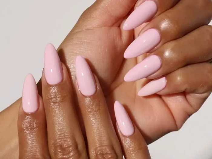 A pair of hands showcasing a simple pink nail design, with long, almond-shaped nails painted in a classic, opaque light pink polish against a white background.