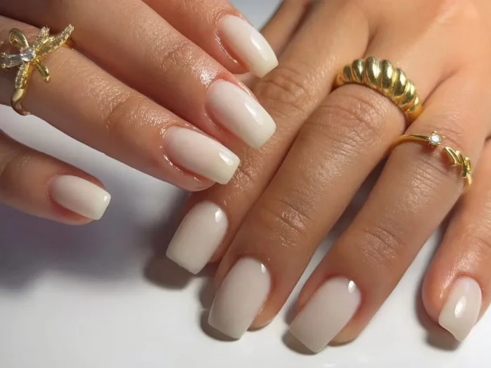 A close-up of hands with an elegant, simple white nail design in a milky finish, adorned with gold rings.