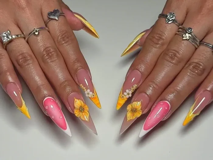Close-up of hands displaying long, vibrant stiletto nails with yellow and orange gradient tips, 3D yellow and white flowers, and pink accent nails with raised white dots.